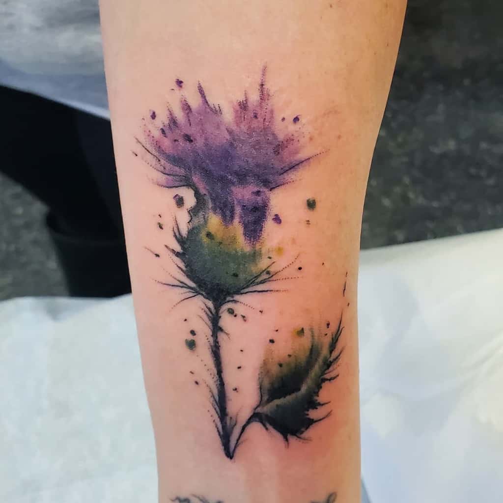 Watercolor-style tattoo of a delicate thistle rendered in soft purples, gracefully adorning a person's forearm
