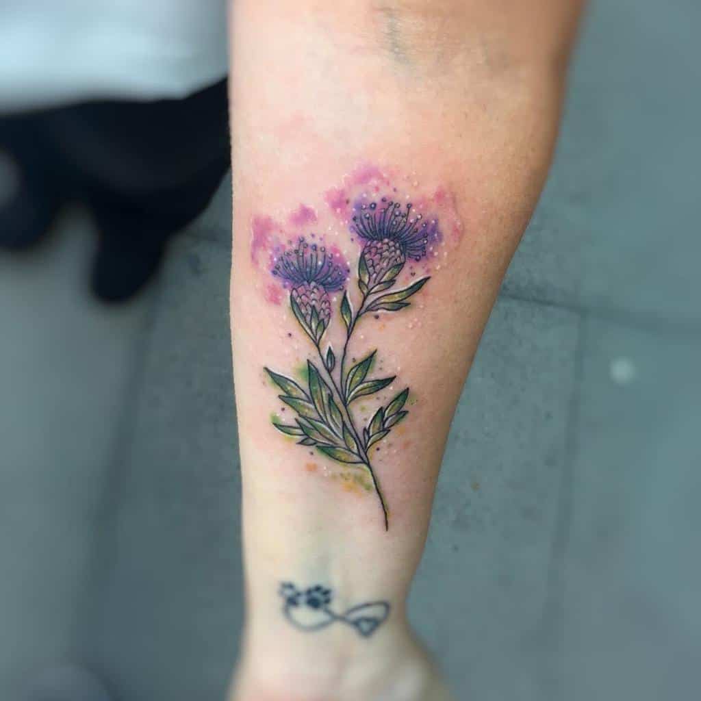 Vibrant forearm tattoo of purple thistle flowers with green leaves, highlighted by pink watercolor accents