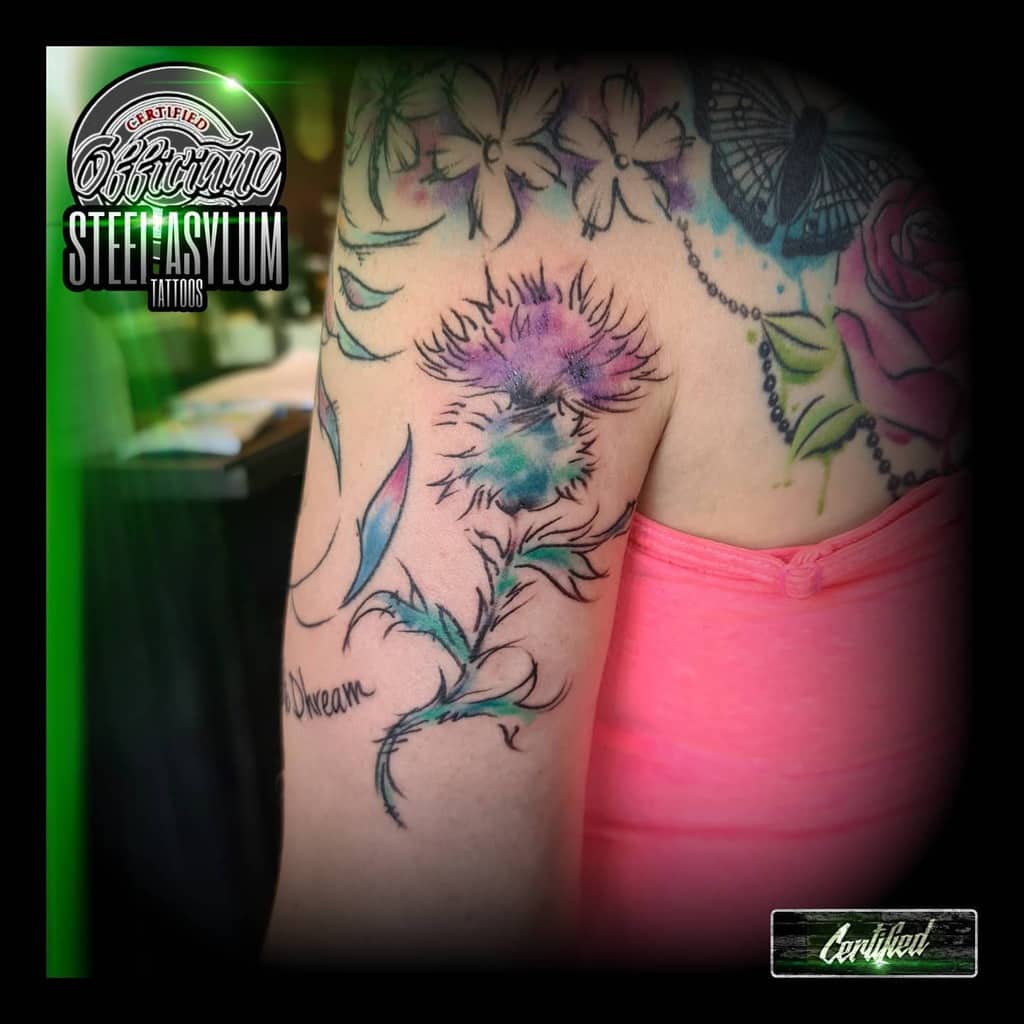 Colorful upper arm tattoo of a thistle with vibrant watercolor splashes in purple, pink, and blue, accented by delicate leaves