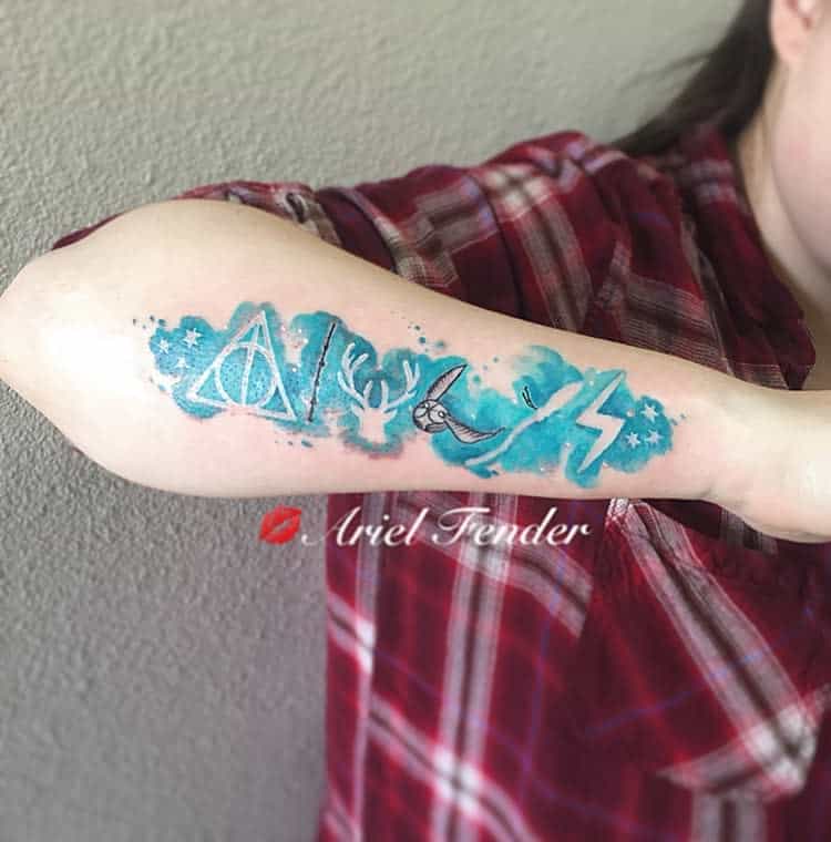 Harry Potter-themed tattoo with vibrant blue watercolor design featuring the Deathly Hallows, a stag, Snitch, and lightning bolt