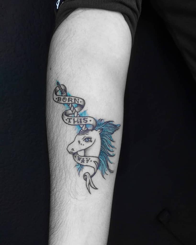 Tattoo of a unicorn with blue mane and a ribbon that reads "Born This Way" on a forearm