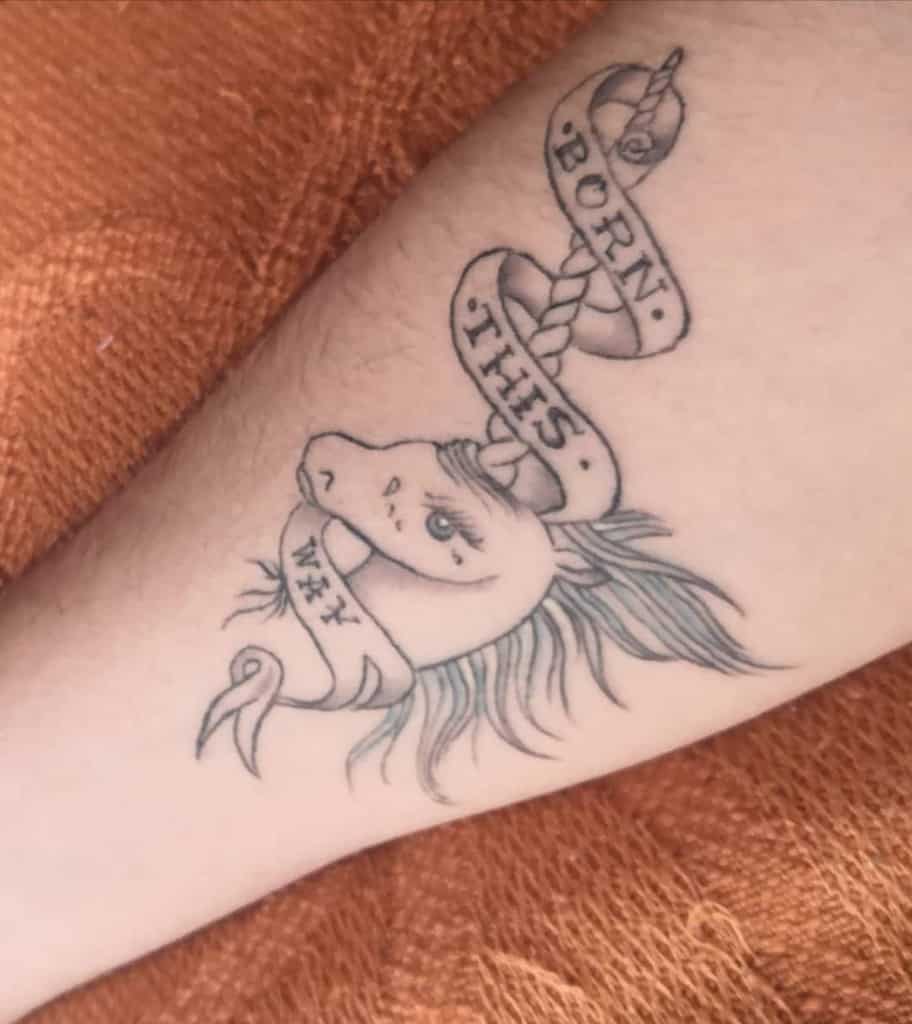 Tattoo of a unicorn head with a flowing mane and a ribbon that reads "Born This Way" on an arm