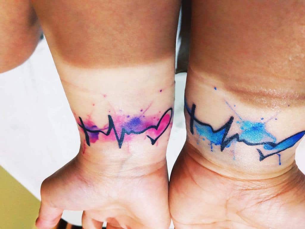 Vibrant watercolor wrist tattoos featuring a cross, heartbeat line, and heart symbol, with pink, purple, and blue splashes