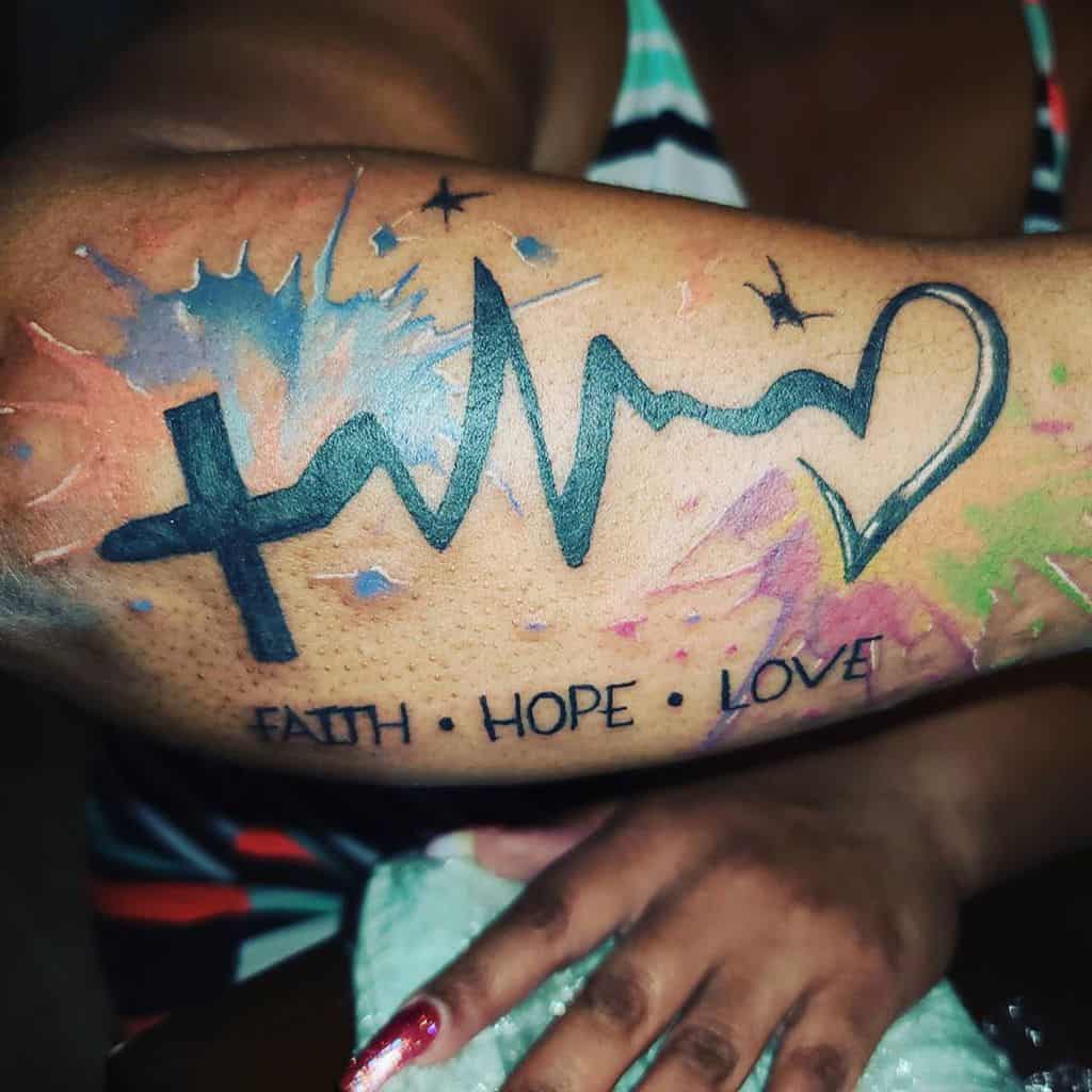 Arm tattoo with a cross, heartbeat, and heart design, featuring colorful splashes and the words "Faith Hope Love"