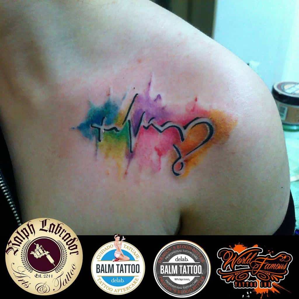 Watercolor tattoo on the shoulder featuring a cross, heartbeat line, and heart symbol, with a vibrant background of blue, green, yellow, and pink hues