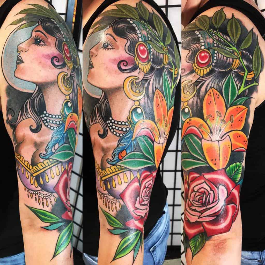 Vibrant arm tattoo of a gypsy woman adorned with jewelry, surrounded by red roses, orange lilies, and lush green leaves