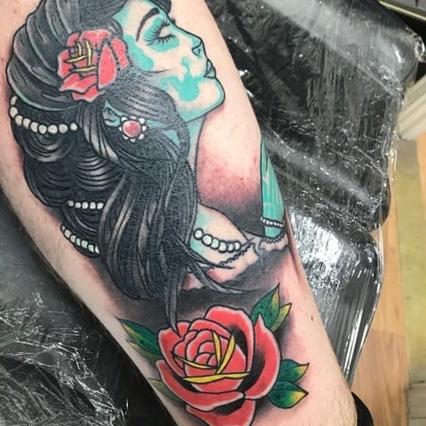 A colorful tattoo of a woman with a skull face, roses in her hair, and a large red rose at the bottom, on the forearm
