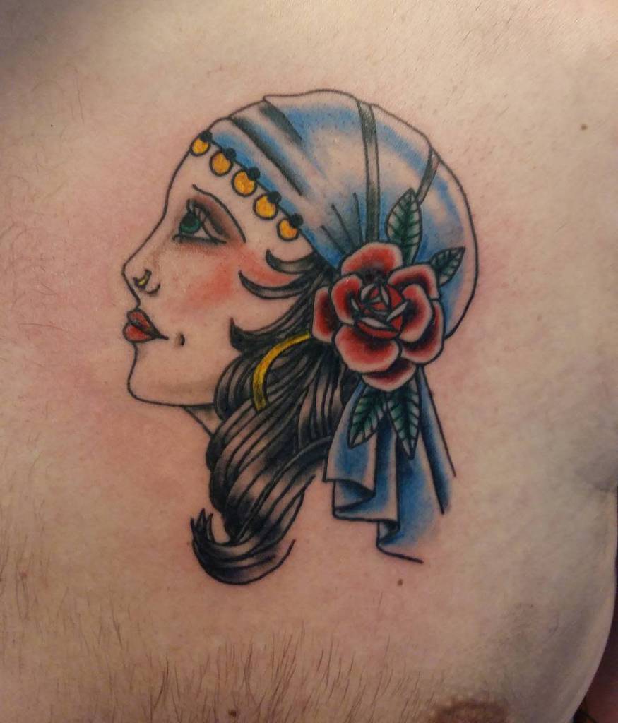 A vibrant tattoo features a gypsy woman adorned in a headscarf gracefully entwined with a rose, beautifully etched on the chest