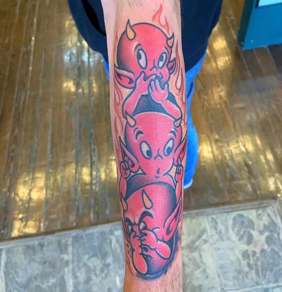 Forearm tattoo of cartoon devils stacked vertically, each covering ears, mouth, and eyes