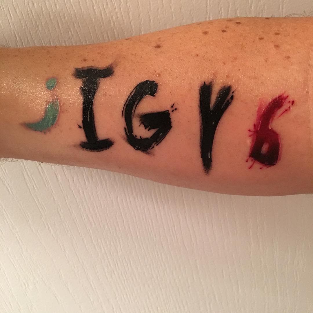 Arm with a colorful tattoo featuring the letters "I G Y 6" with a semicolon in teal and red accents on the "6"