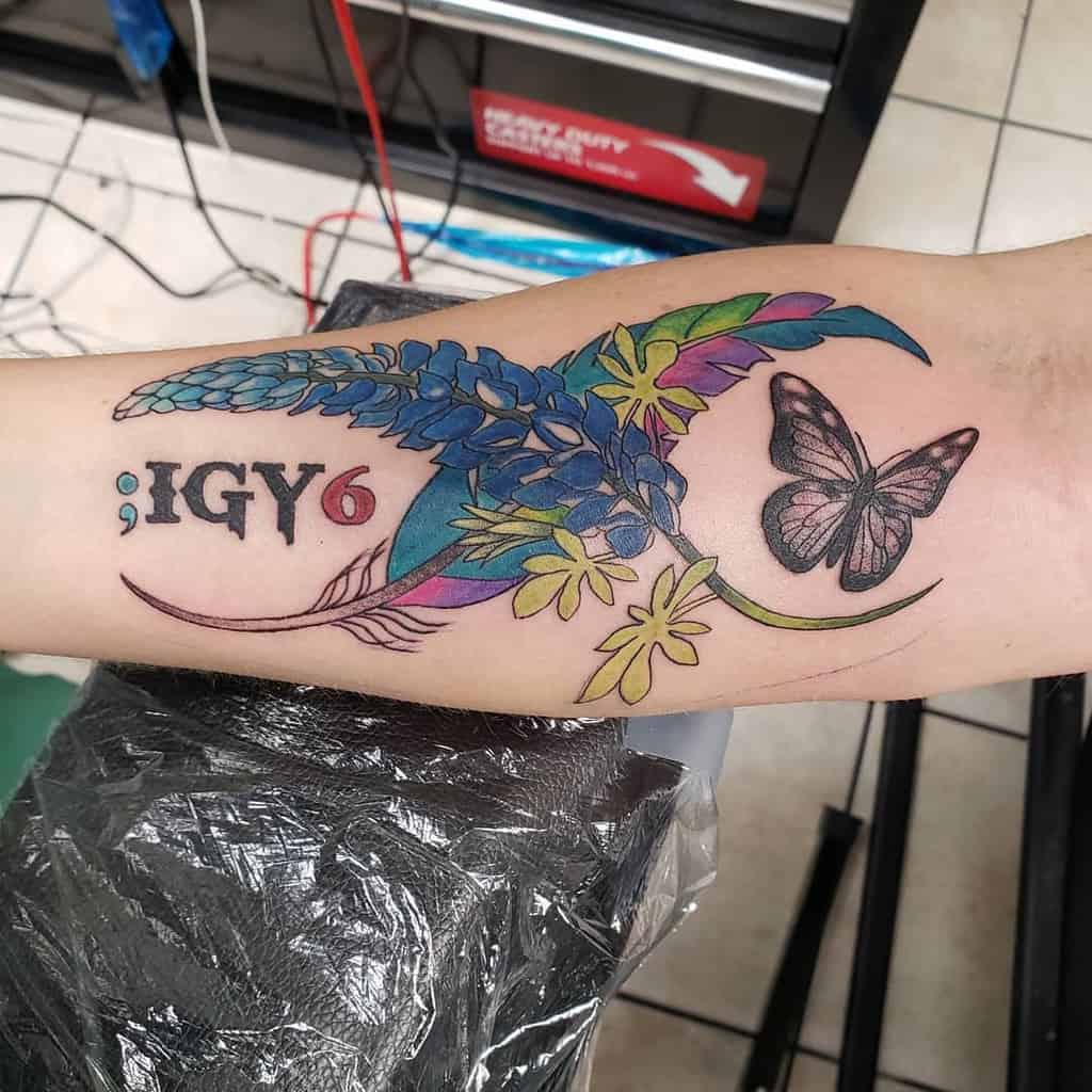 Arm tattoo featuring flowers, a butterfly, and the text "; IGY6" in black ink, with colorful accents on a light skin tone