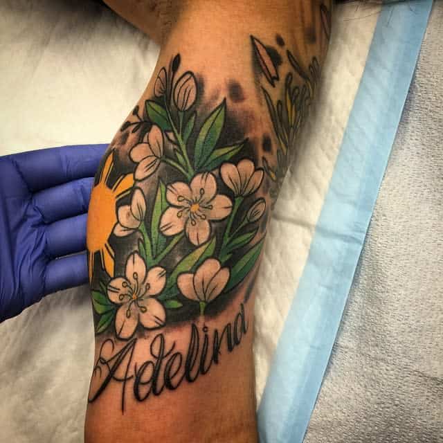 Colorful tattoo of jasmine flowers, leaves, and a sun with the name "Adelina" on an arm