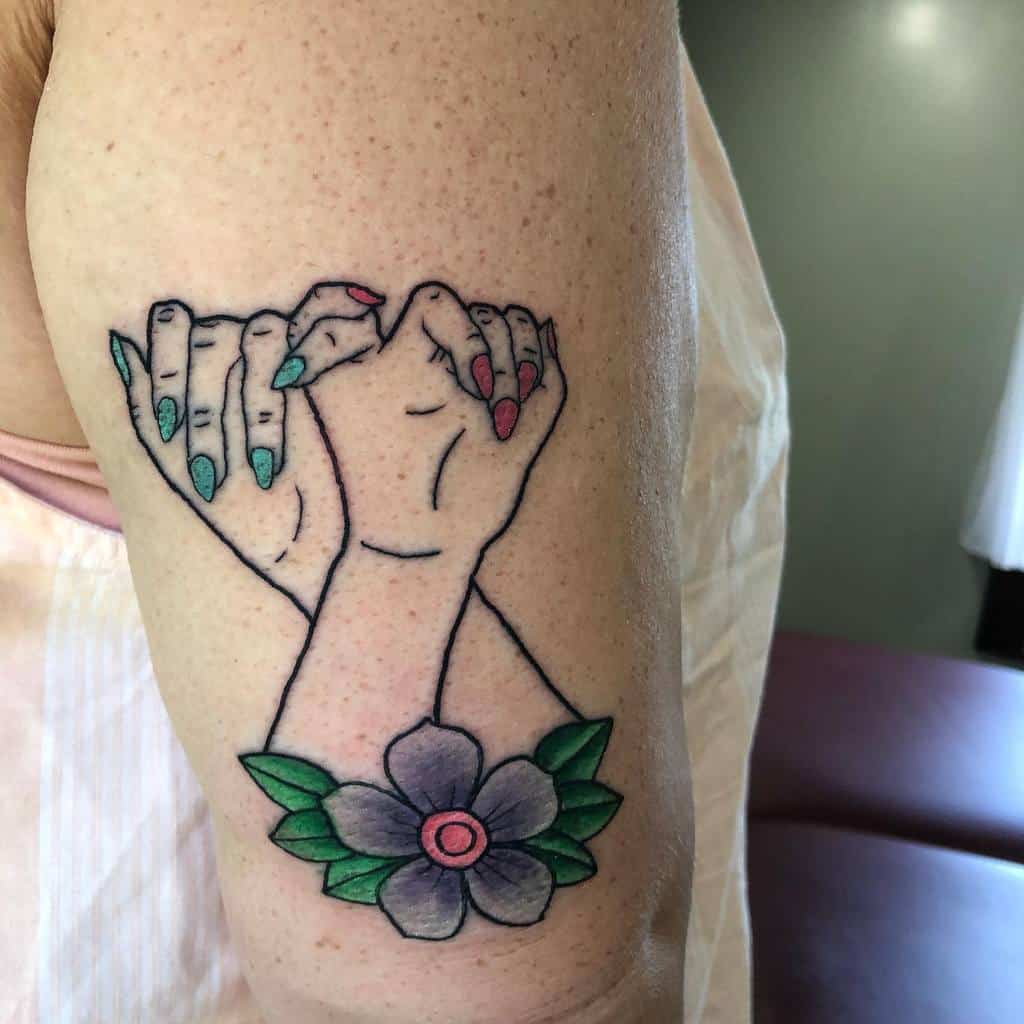 Tattoo of two hands making a pinky promise above a flower with green leaves and a pink center on an arm
