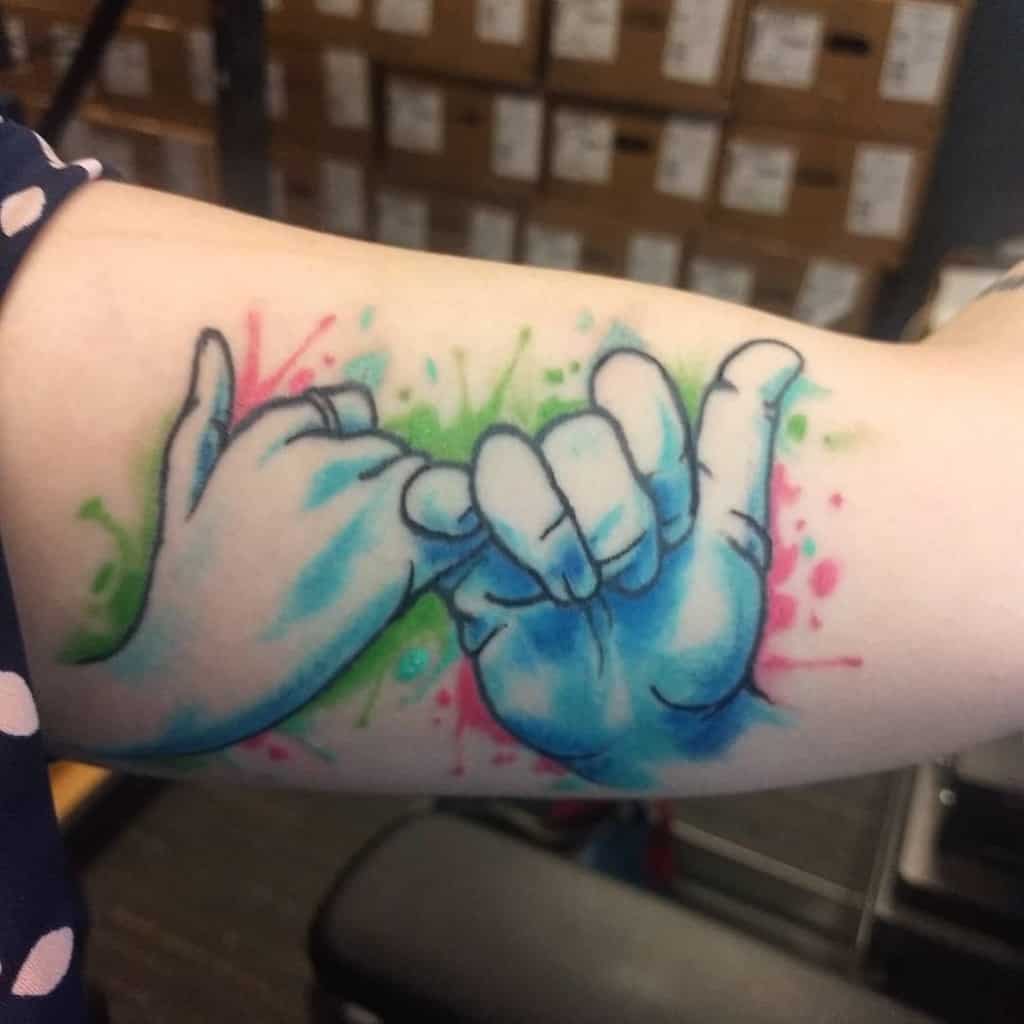 Tattoo of two hands making a pinky promise, with colorful splashes of green and pink in the background
