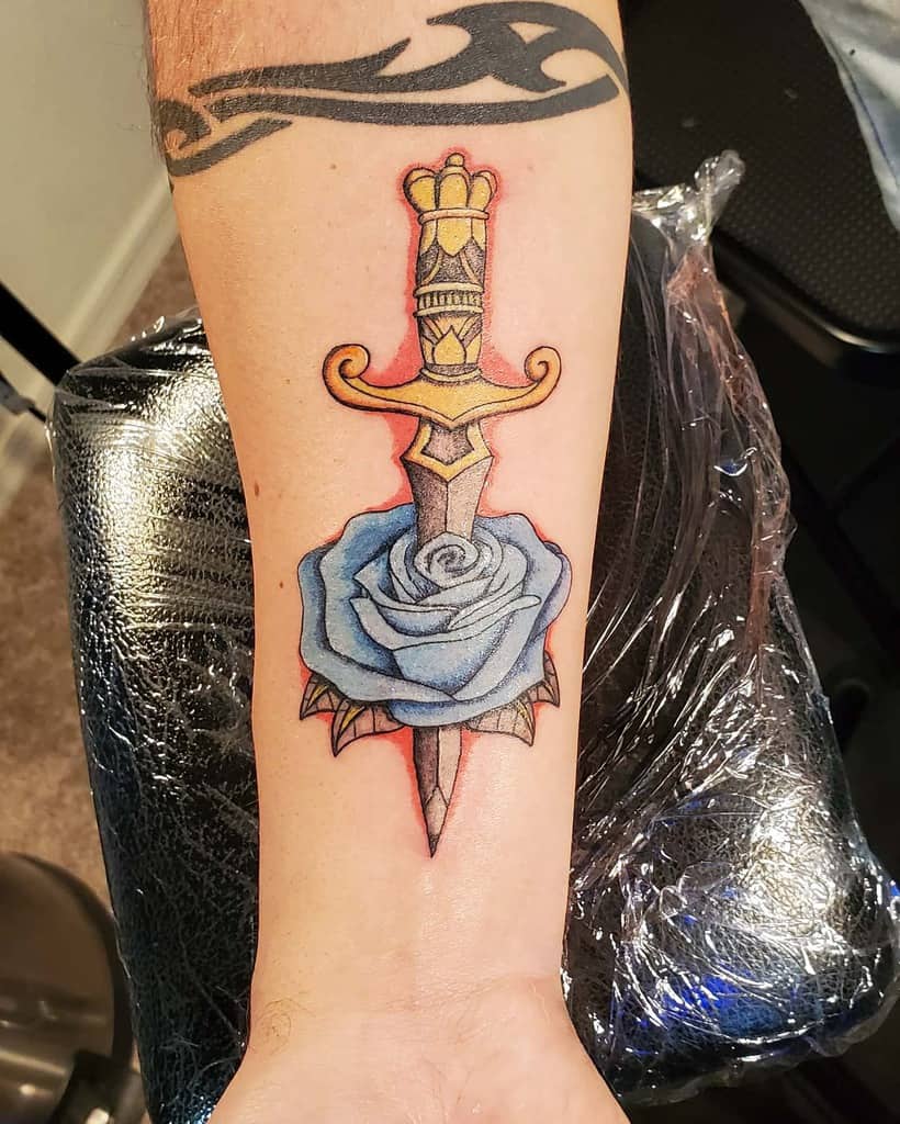 Tattoo of a dagger piercing a blue rose on a forearm with a tribal band tattoo