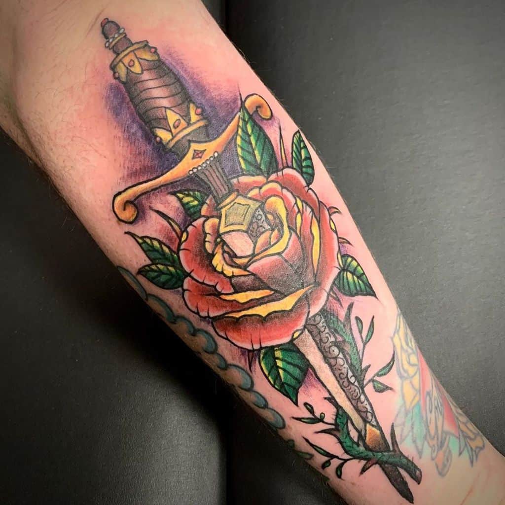 Tattoo of a dagger piercing a red rose with green leaves on an arm