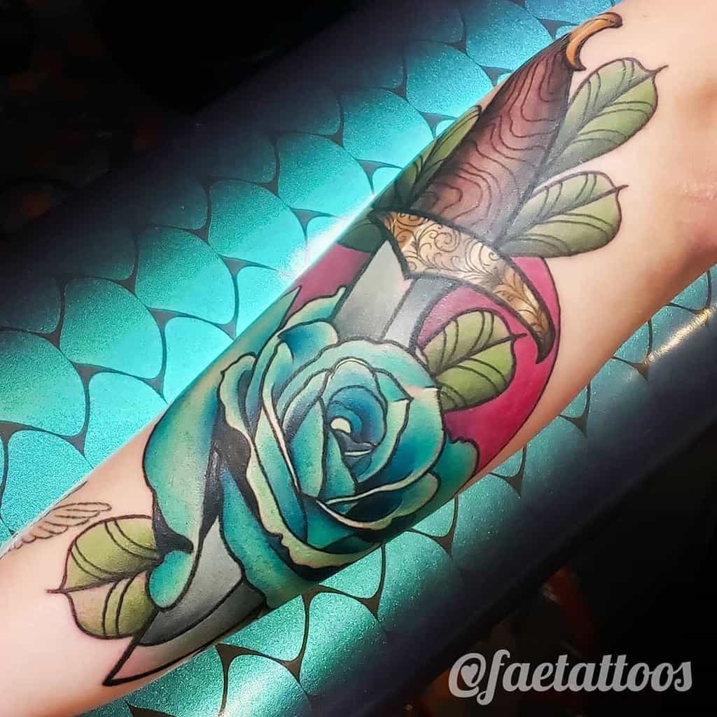 Tattoo of a dagger with a blue rose on a forearm