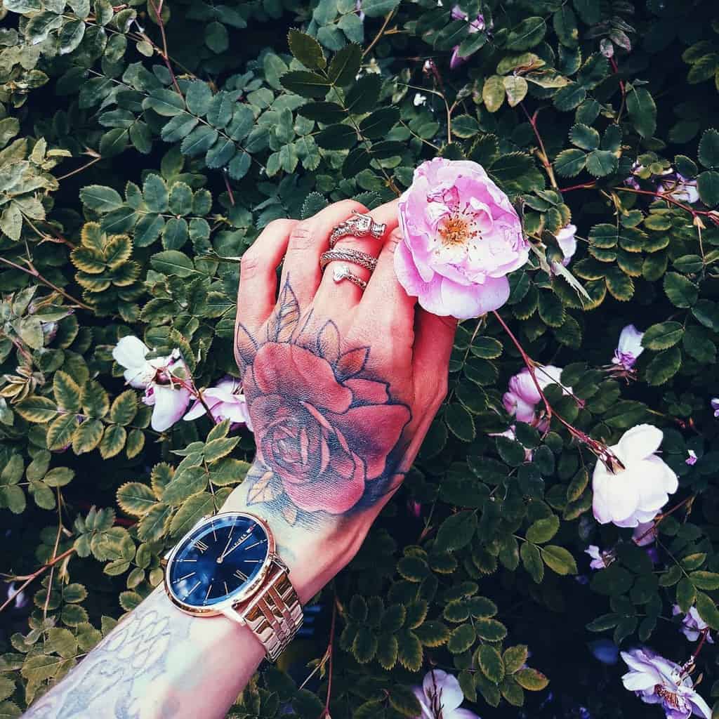 A vibrant watercolor-style tattoo of a rose on the hand, surrounded by rings, a watch, and blooming flowers in the background