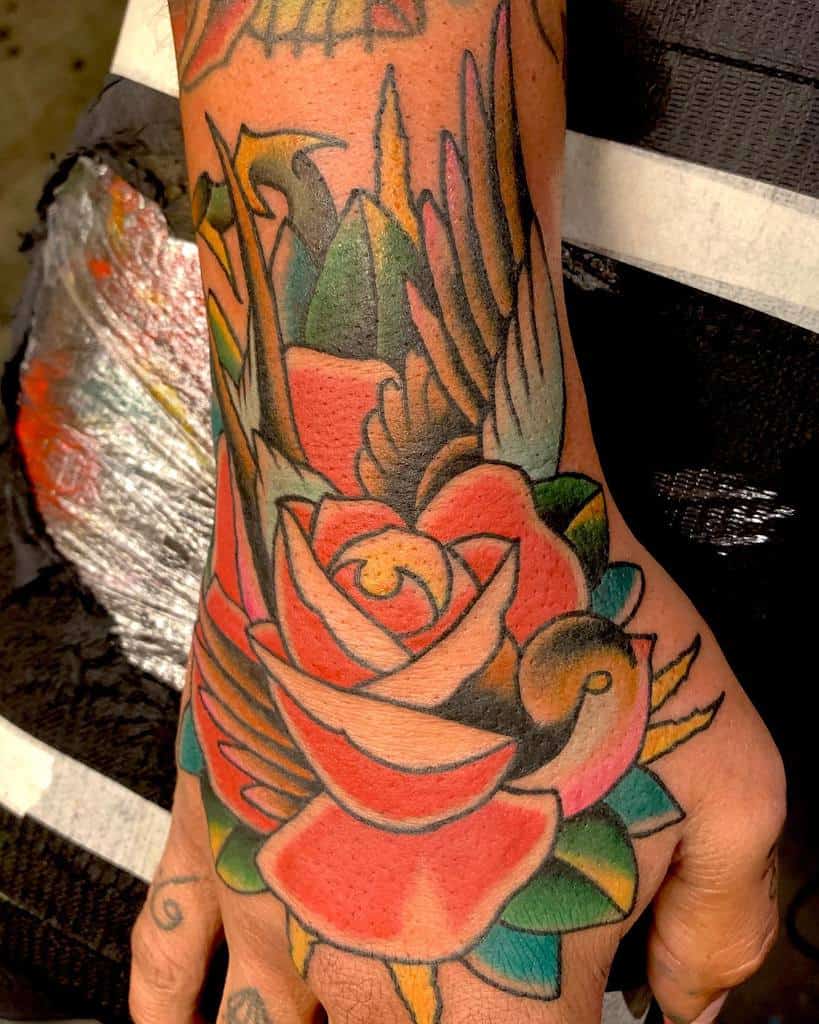 A colorful traditional-style rose tattoo on the hand, featuring bold outlines, vibrant red petals, green leaves, and bird accents, creating a dynamic and lively design