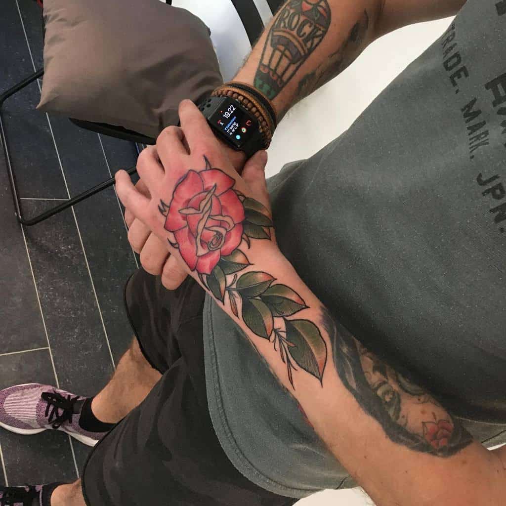 A colorful rose tattoo on the hand and forearm, featuring vibrant red petals, green leaves, and bold outlines
