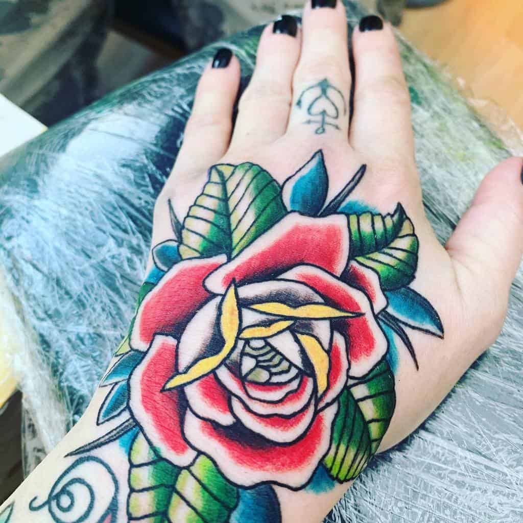 A colorful traditional rose tattoo on the hand, featuring bold red petals, yellow accents, and green leaves, creating a vibrant and eye-catching design