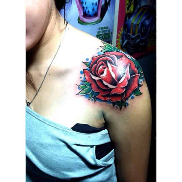 A vibrant red rose with lush green leaves is elegantly inked on the shoulder