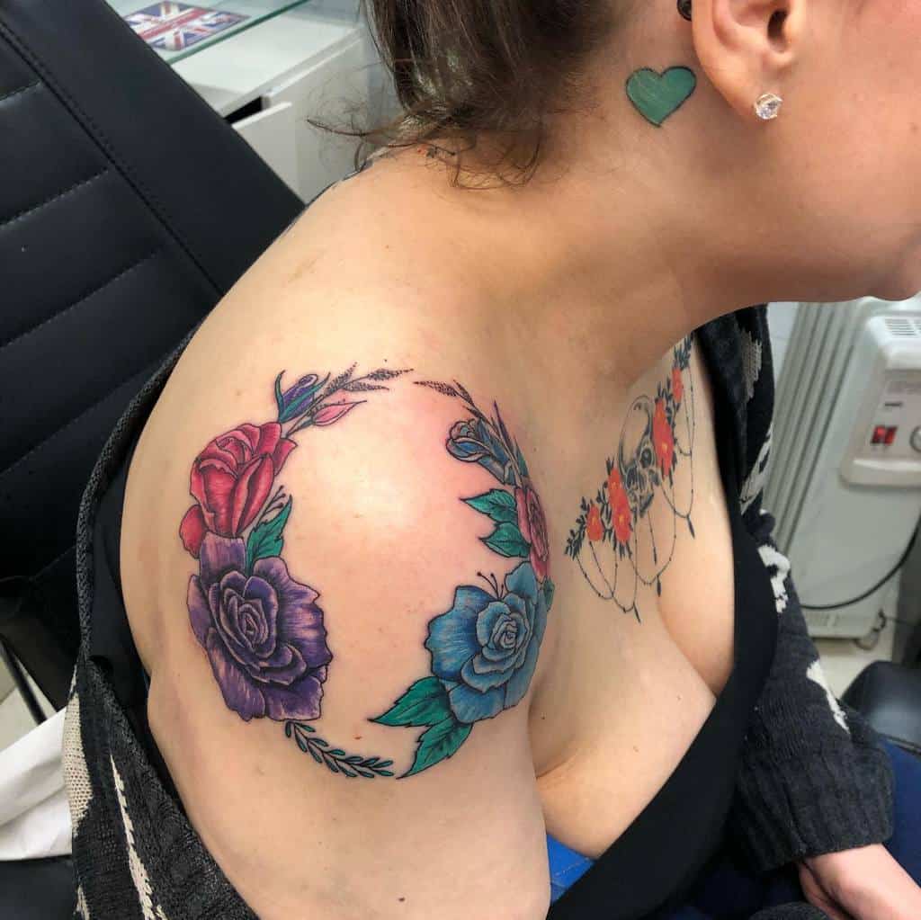 A colorful watercolor rose tattoo on the shoulder, featuring red, purple, and blue roses with vibrant leaves and soft blending of colors
