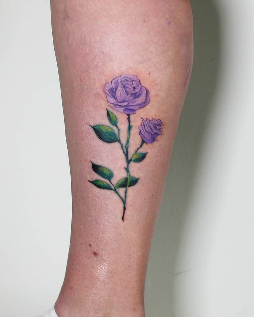 A tattoo of purple roses with lush green leaves and a delicate stem spiraling down a leg