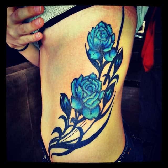 A vibrant tattoo of blue roses with swirling black tribal patterns, placed along the side of the torso, with a watercolor effect
