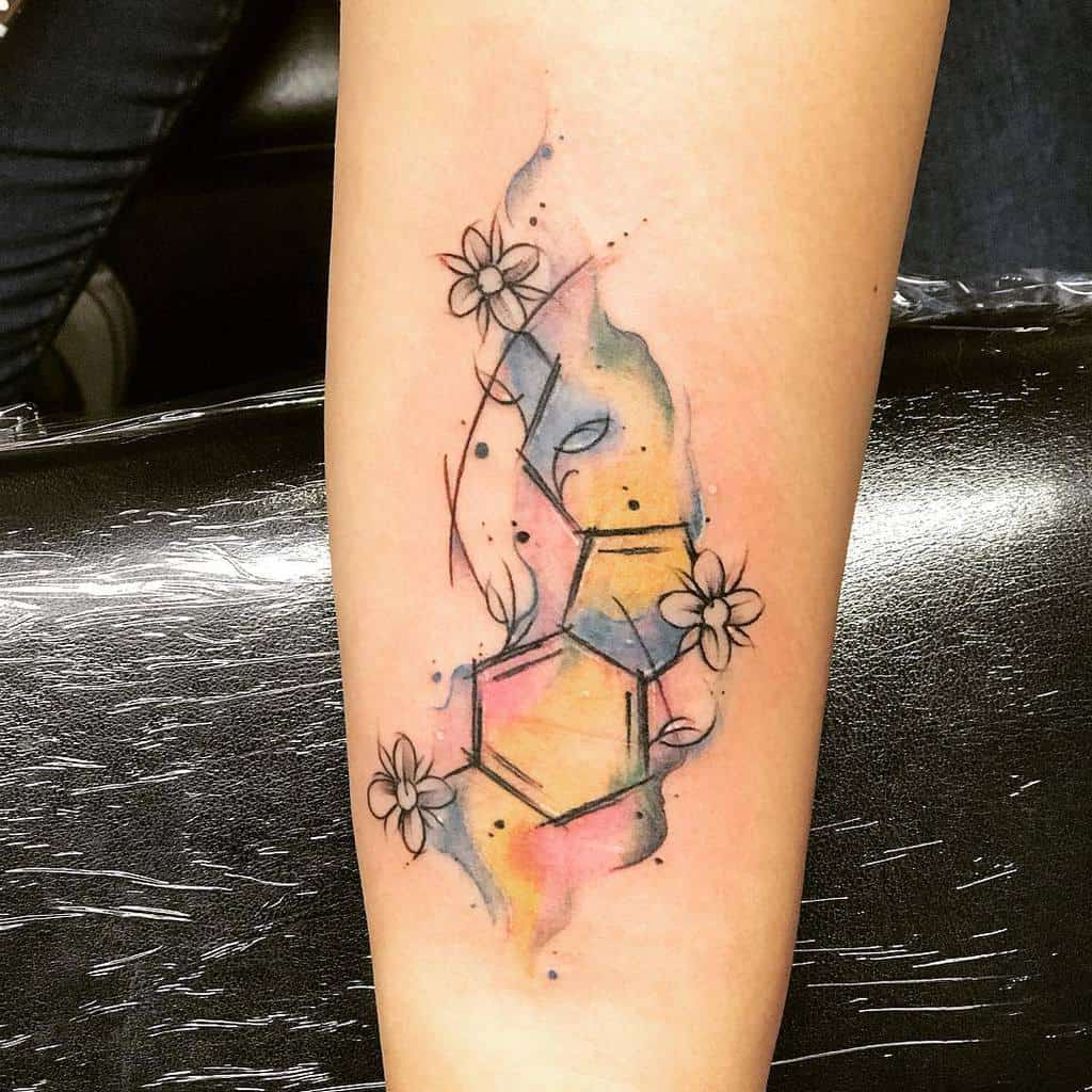 Watercolor-style tattoo of the serotonin molecule, surrounded by flowers and abstract splashes of pink, blue, and yellow