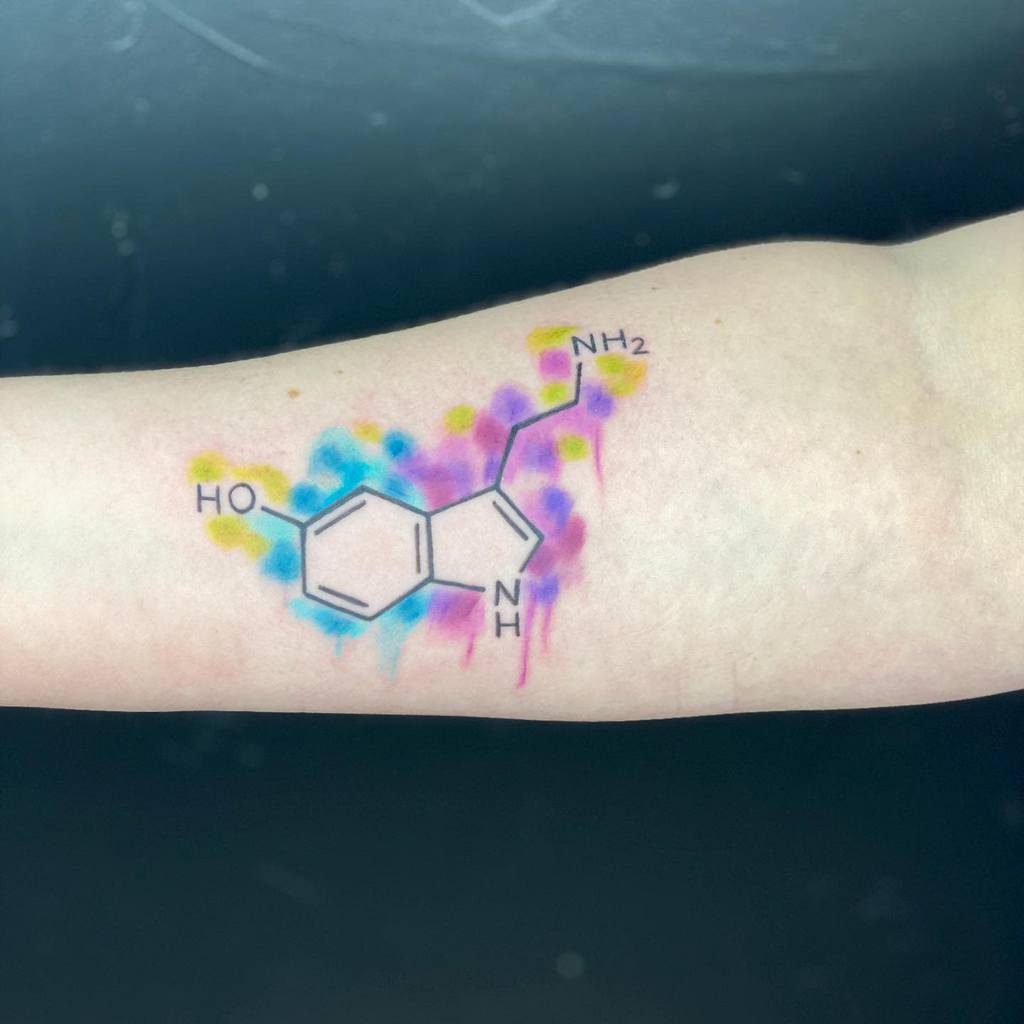 Serotonin molecule tattoo with vibrant watercolor splashes in blue, pink, and yellow on the forearm