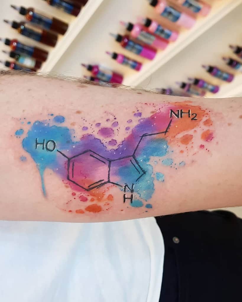 Watercolor-style serotonin tattoo on an arm with vibrant splashes of blue, pink, and purple