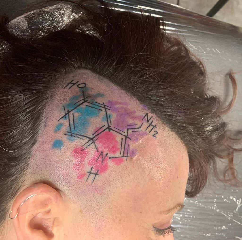 Serotonin molecule tattoo on the side of the head with blue and pink watercolor accents under shaved hair