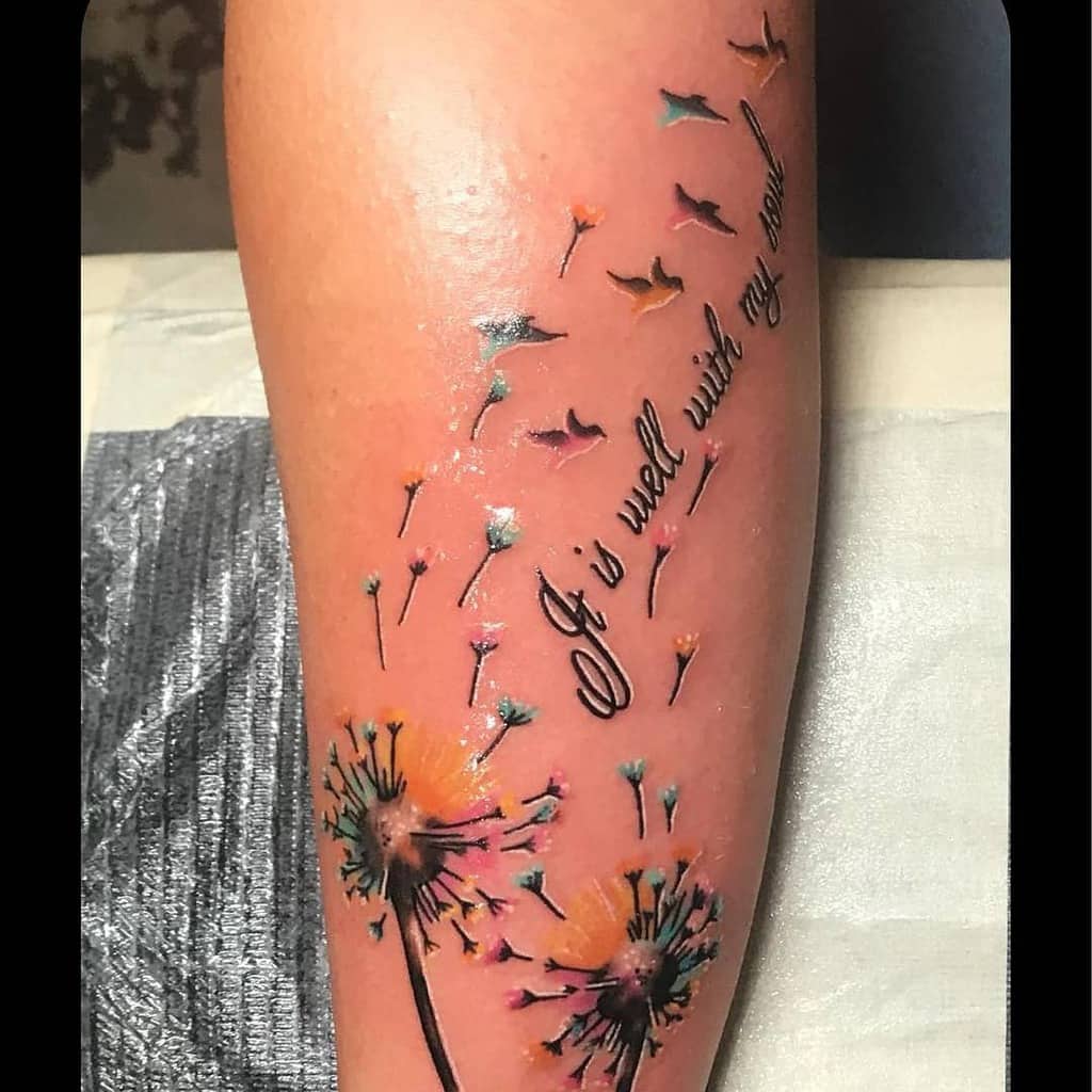 A vibrant tattoo featuring dandelions with seeds blowing off, accompanied by the quote "It is well with my soul" in flowing script.