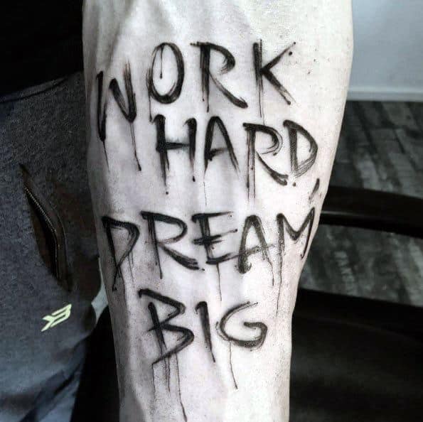 Forearm tattoo with the phrase “Work Hard Dream Big” in bold, brushstroke-style letters with dripping ink effect for a raw, artistic look.