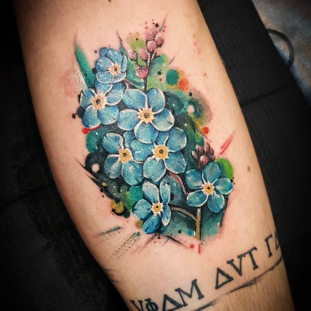 A tattoo of delicate blue forget-me-nots intertwined with leaves adorns the arm, beautifully complementing a nearby Greek script tattoo