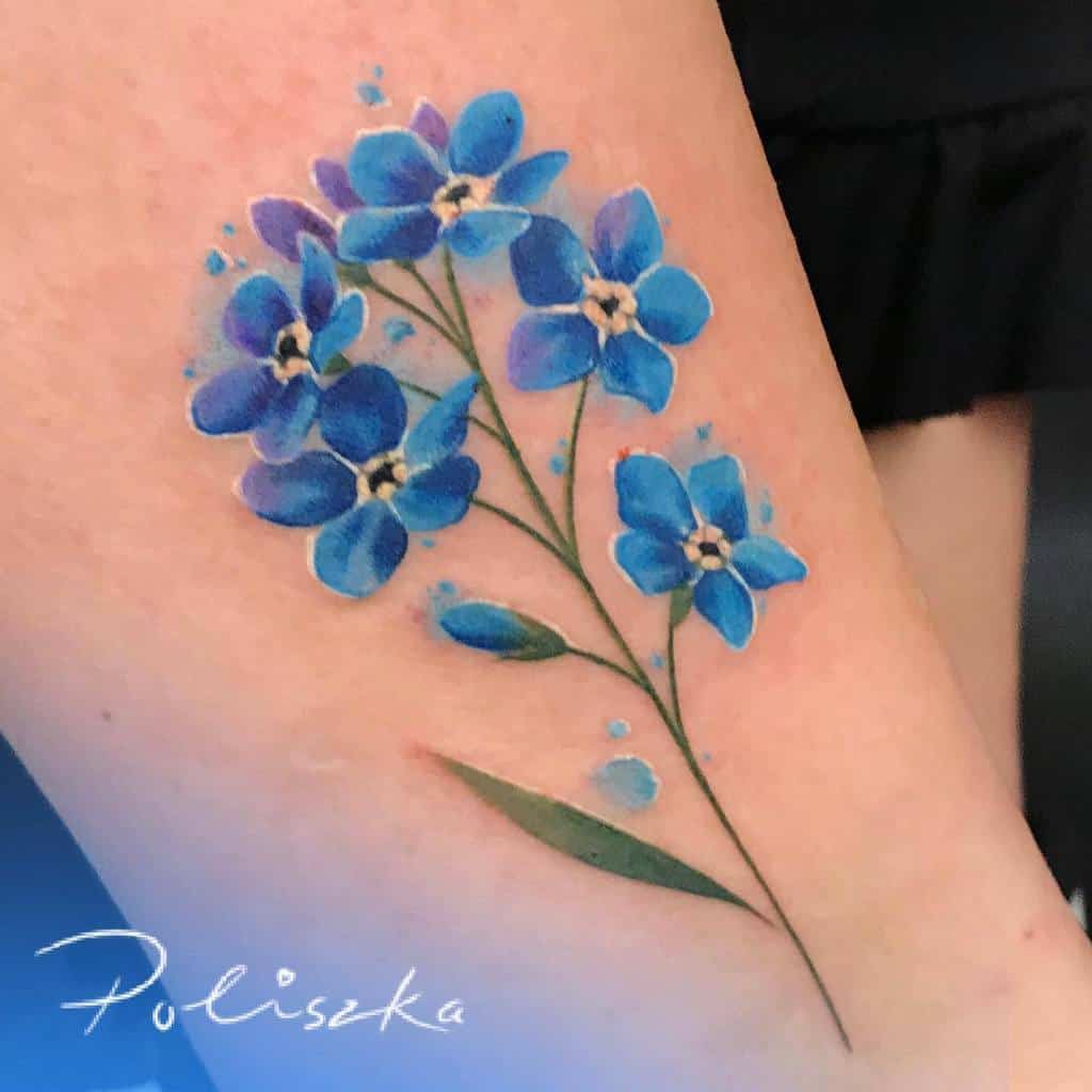 A delicate watercolor tattoo of forget-me-nots in shades of blue with soft green leaves, beautifully inked on the arm