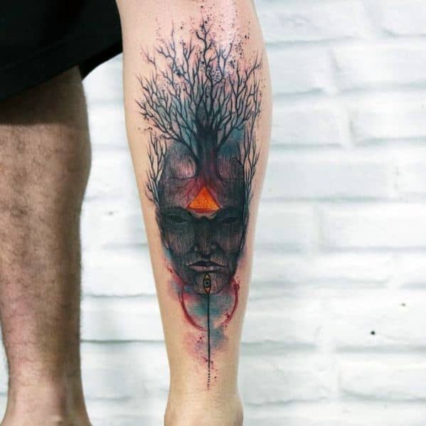 Surreal tree tattoo on the leg, featuring a human face merging with branches, a glowing orange triangle, and abstract watercolor effects.
