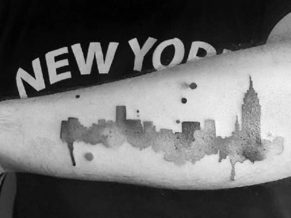 Watercolor-style tattoo of the New York City skyline on the forearm, featuring ink splashes and the Empire State Building.