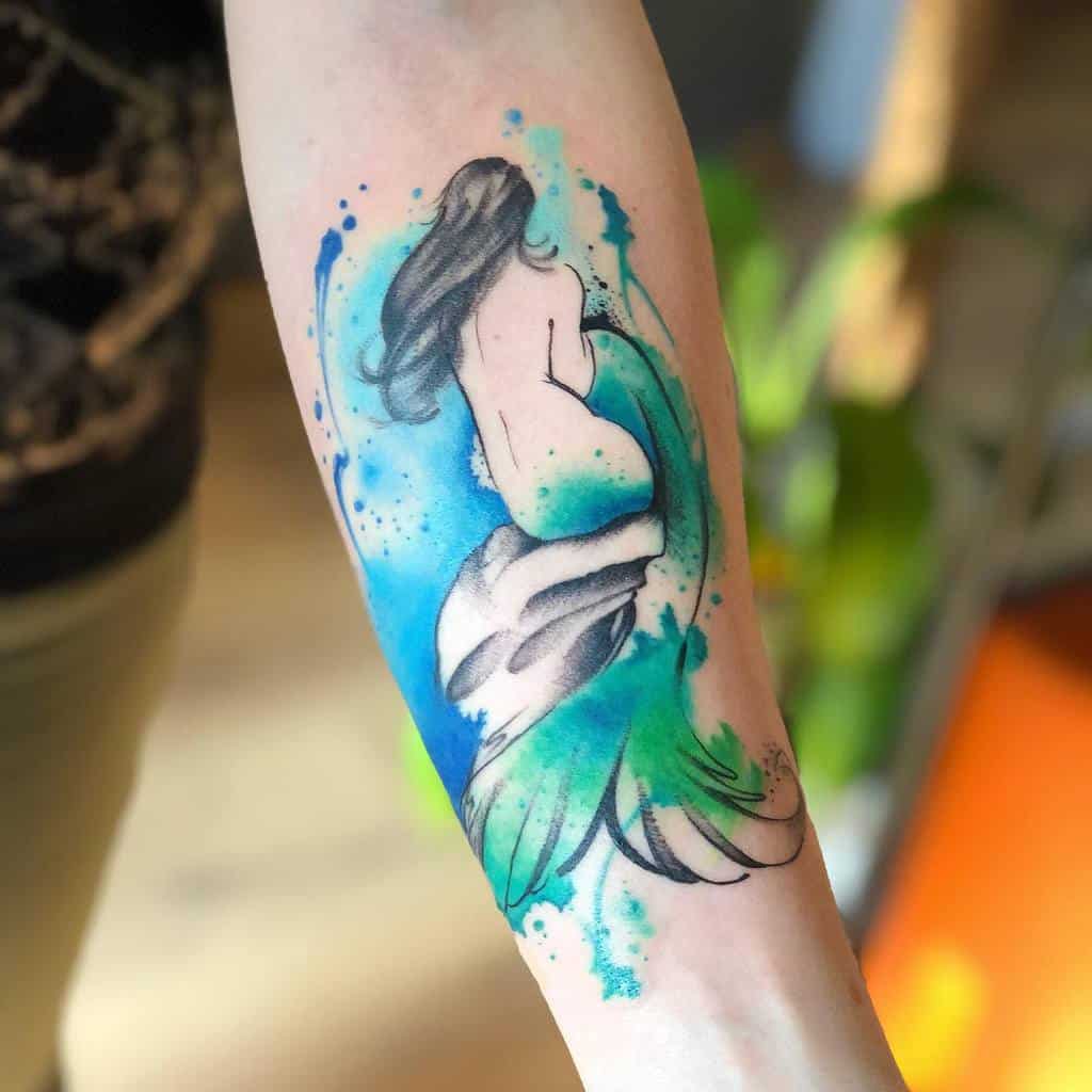 Watercolor-style tattoo of a mermaid sitting on a rock, with flowing hair and a green tail, surrounded by vibrant blue splashes.