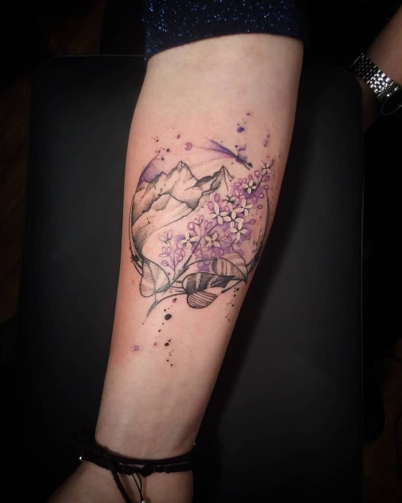 Artistic forearm tattoo featuring lilac flowers, a mountain landscape, and watercolor-style splashes in purple and black ink.