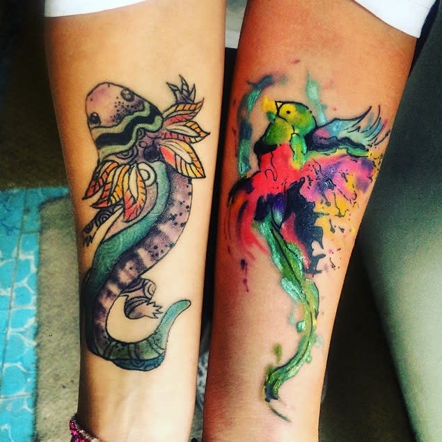 Watercolor-style quetzal tattoo on the forearm, featuring a vibrant splash of green, red, yellow, and purple blending into the bird's body and flowing tail feathers