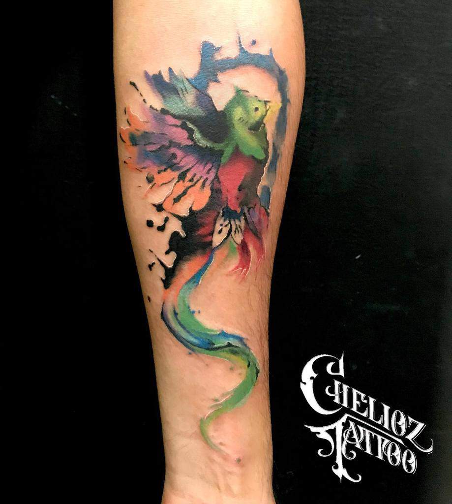 Watercolor-style quetzal tattoo on the forearm, featuring a vibrant green and red bird in flight with abstract splashes of blue, purple, orange, and black ink blending into its wings and tail