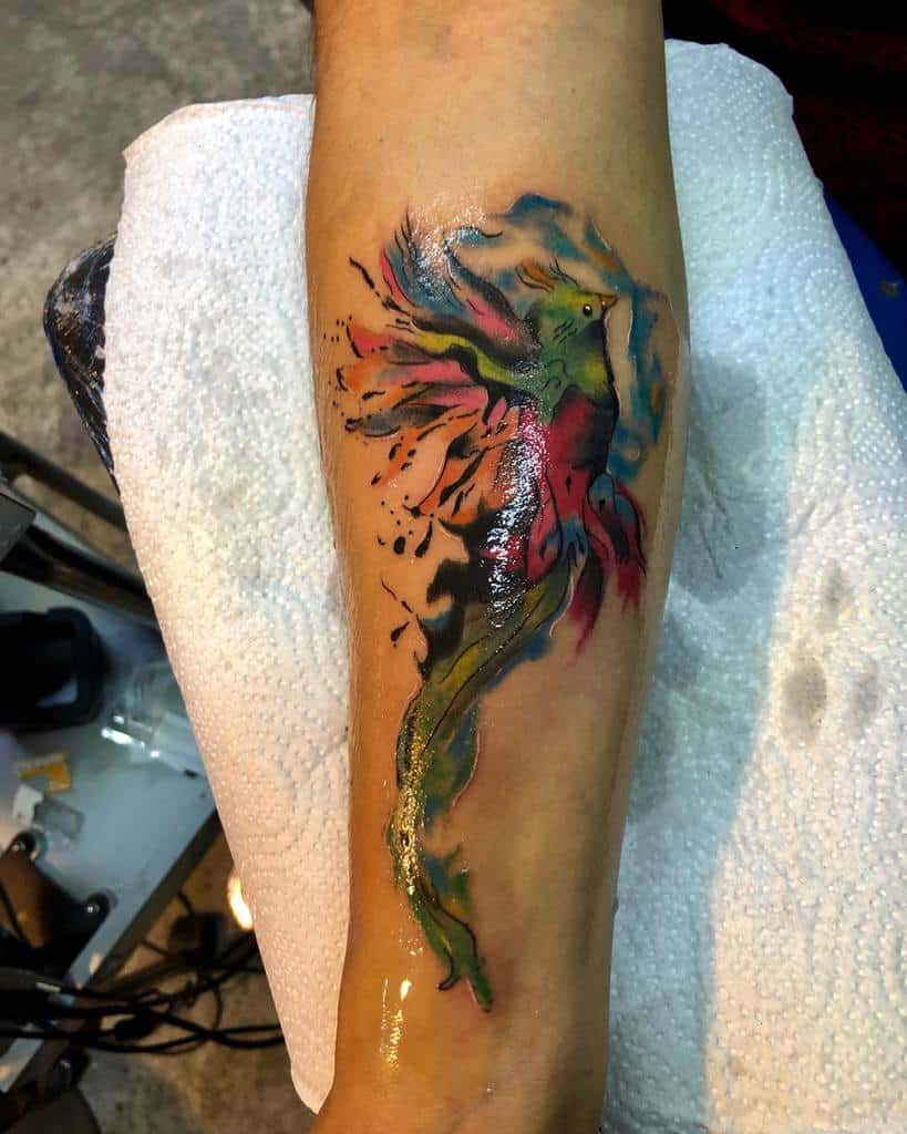 Watercolor-style quetzal tattoo on the forearm, featuring a vibrant green and red bird in flight with abstract splashes of blue, orange, and black ink blending into its wings and flowing tail feathers