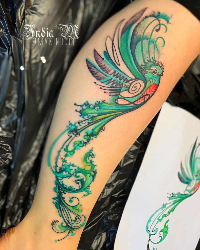 Artistic watercolor-style quetzal tattoo on the leg, featuring a stylized green and red bird in flight with intricate swirling tail feathers and abstract blue-green ink splashes
