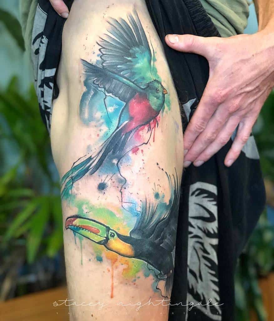 Watercolor-style tattoo on the thigh, featuring a vibrant quetzal in flight with splashes of blue, red, and green ink, accompanied by a colorful toucan below, blending into an abstract background