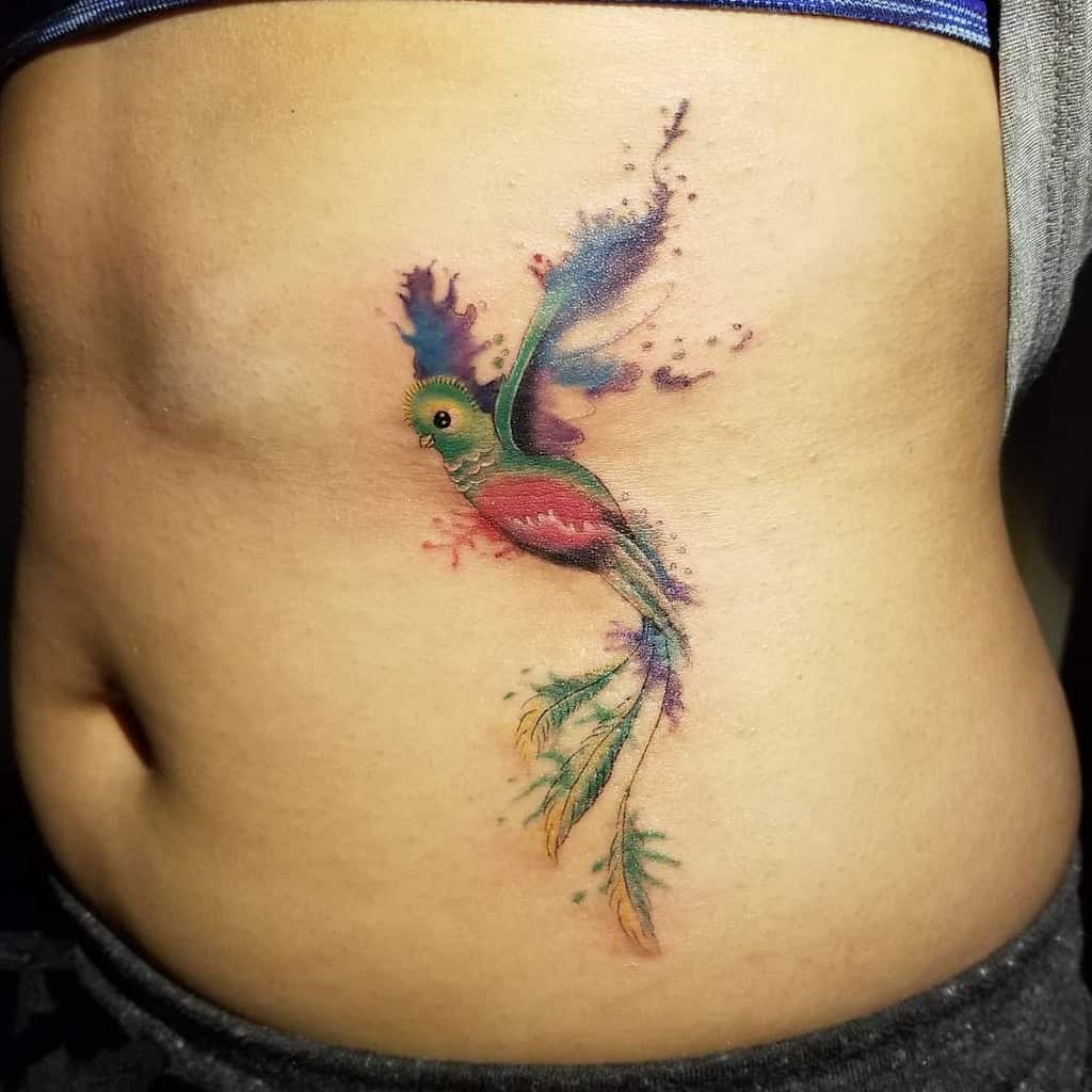 Quetzal-inspired tattoo on a person's side, with blue, pink, and green hues in a watercolor style and flowing tail feathers