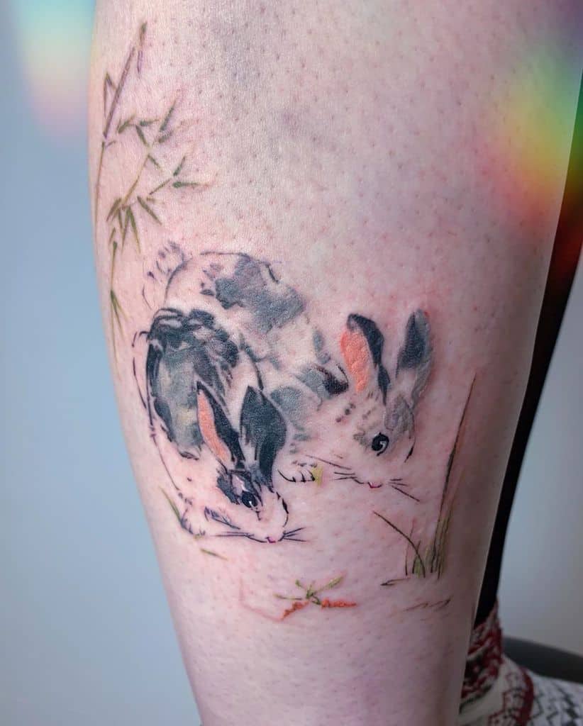 Realistic tattoo of two rabbits grazing in a grassy area, surrounded by delicate bamboo and soft shading
