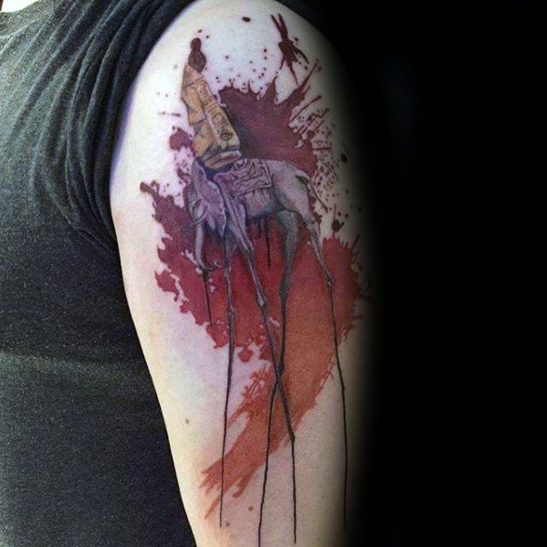 Salvador Dali-inspired upper arm tattoo of a surreal elephant with spindly legs and an obelisk, set against a bold red watercolor splash.