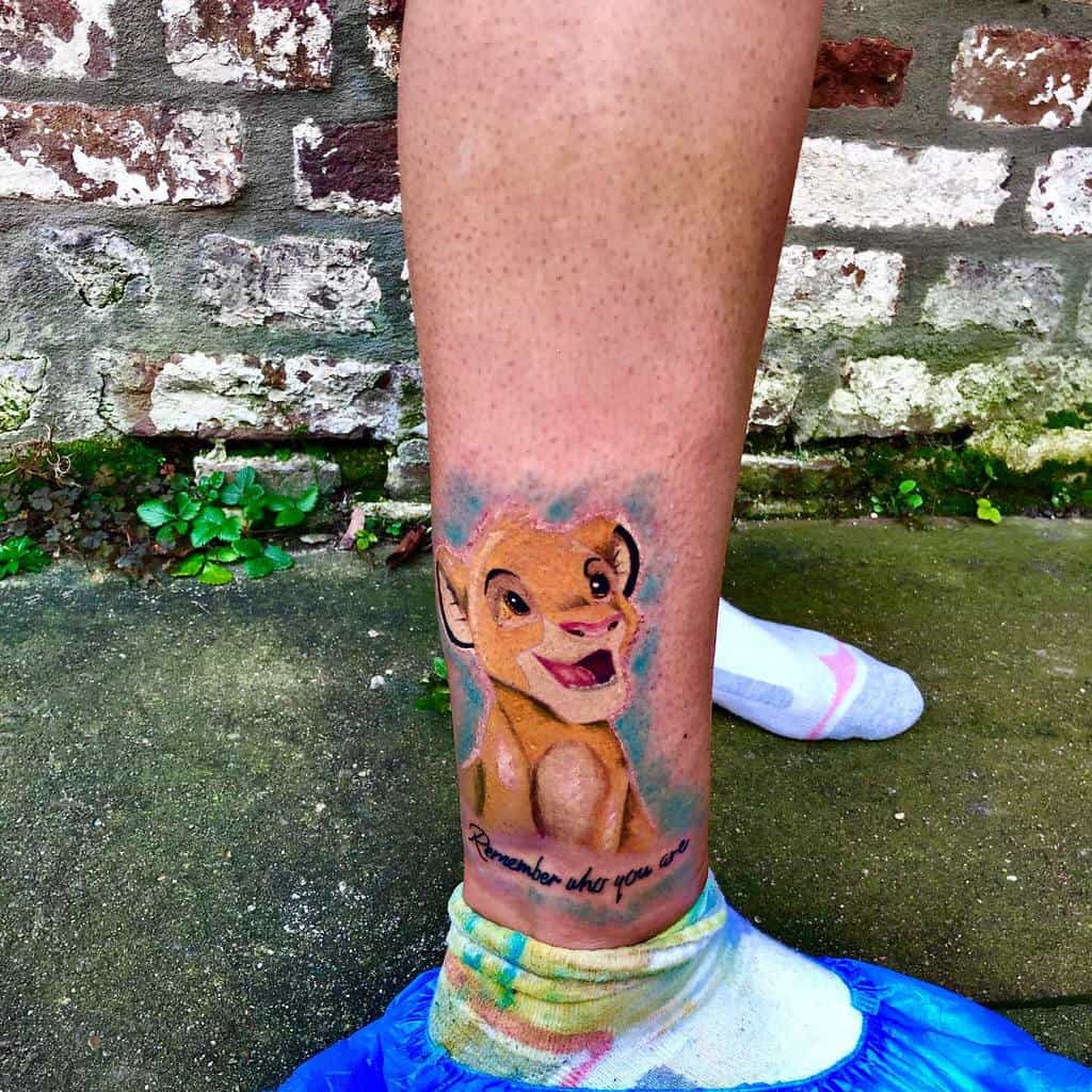 Vivid tattoo of young Simba smiling with the inspiring phrase 'Remember who you are' below, surrounded by soft, colorful shading